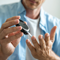 A man in a light blue shirt holds a blood glucose meter in one hand and a test strip over the fingertips of his other hand, ready to take a finger-prick reading.