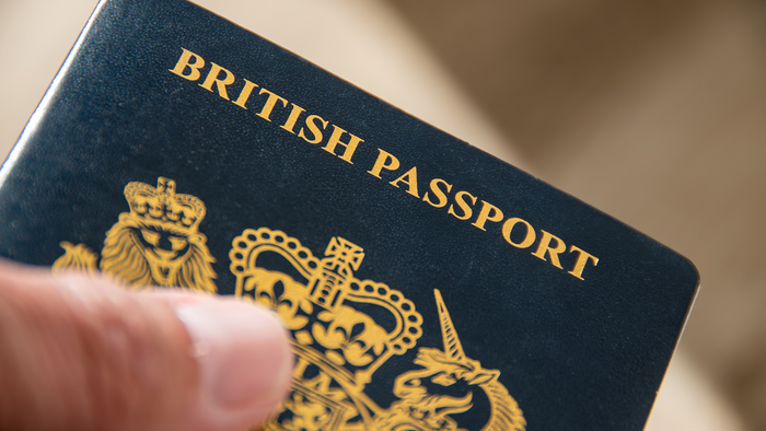 A hand holding a dark blue British passport, with gold lettering and the royal coat of arms on the cover