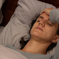 A woman lying in bed with her eyes closed and one hand pressed to her forehead, wearing a grey long-sleeved top against grey pillows.