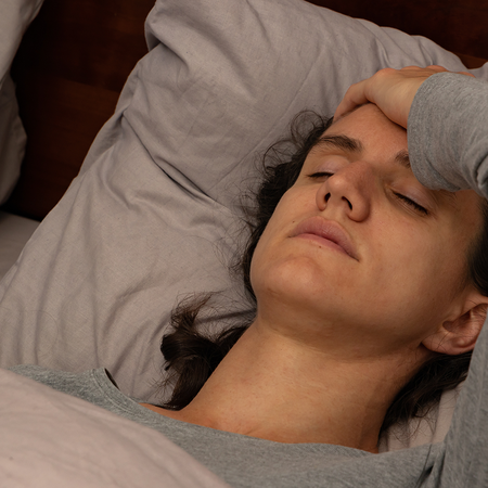 A woman lying in bed with her eyes closed and one hand pressed to her forehead, wearing a grey long-sleeved top against grey pillows.