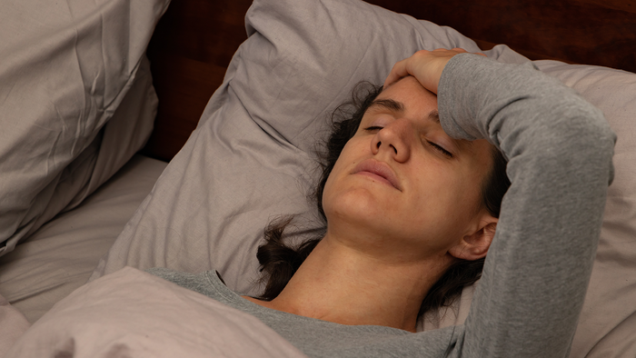 A woman lying in bed with her eyes closed and one hand pressed to her forehead, wearing a grey long-sleeved top against grey pillows.