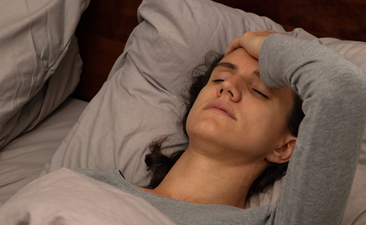A woman lying in bed with her eyes closed and one hand pressed to her forehead, wearing a grey long-sleeved top against grey pillows.