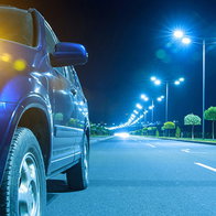 Car with headlights on beside a brightly lit road at night.