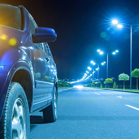 Car with headlights on beside a brightly lit road at night.