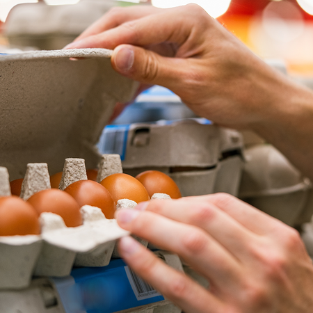 A shopper’s hands lifting the lid of a cardboard egg box to look at the brown eggs inside, on a supermarket shelf of egg boxes.