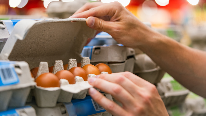 A shopper’s hands lifting the lid of a cardboard egg box to look at the brown eggs inside, on a supermarket shelf of egg boxes.