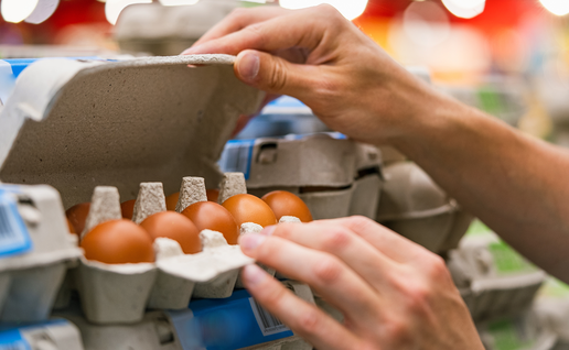 A shopper’s hands lifting the lid of a cardboard egg box to look at the brown eggs inside, on a supermarket shelf of egg boxes.