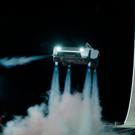 White Tesla Roadster hovering above the ground at night, with plumes of white vapour from thrusters under the car and a tall curved concrete ramp lit with white lines on the right.