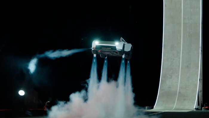 White Tesla Roadster hovering above the ground at night, with plumes of white vapour from thrusters under the car and a tall curved concrete ramp lit with white lines on the right.