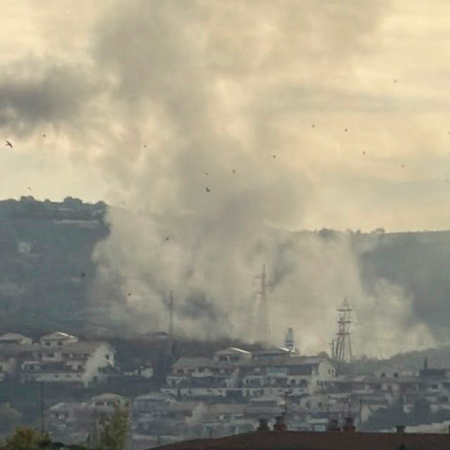 A tall column of grey smoke with a rounded, mushroom-shaped top rises above a hillside of white houses, with trees and a red-brick apartment block in the foreground.