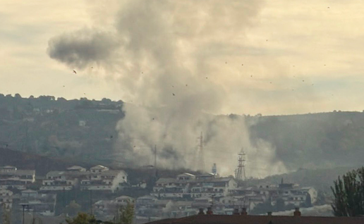 A tall column of grey smoke with a rounded, mushroom-shaped top rises above a hillside of white houses, with trees and a red-brick apartment block in the foreground.