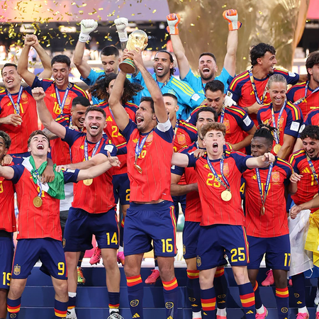 Spain’s football team celebrating on a podium in red shirts and winners’ medals as the captain lifts the World Cup trophy and gold confetti falls.