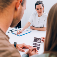 A man and a woman, seen from behind, sit at a white table facing a doctor in a white tunic who is writing on a clipboard, with a stethoscope and three ultrasound scan printouts on the table.
