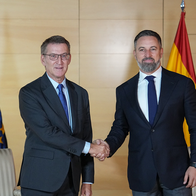 Two men in dark suits shake hands in front of Spanish and European Union flags, with two cream striped armchairs behind them.