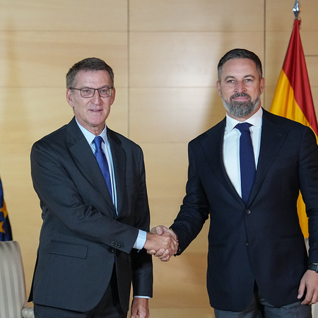 Two men in dark suits shake hands in front of Spanish and European Union flags, with two cream striped armchairs behind them.