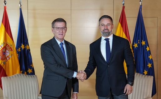 Two men in dark suits shake hands in front of Spanish and European Union flags, with two cream striped armchairs behind them.