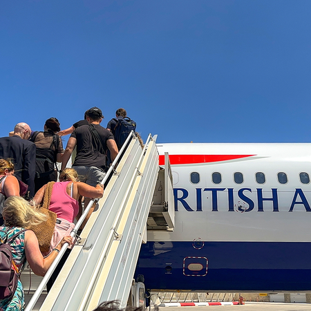 Passengers climb a set of mobile boarding stairs towards a British Airways plane parked on the airport apron under a clear blue sky.