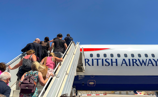 Passengers climb a set of mobile boarding stairs towards a British Airways plane parked on the airport apron under a clear blue sky.