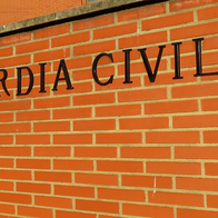 The words “Guardia Civil” in black letters on a red brick wall at the entrance to a brick building, with a raised barrier and street lamps to the left.