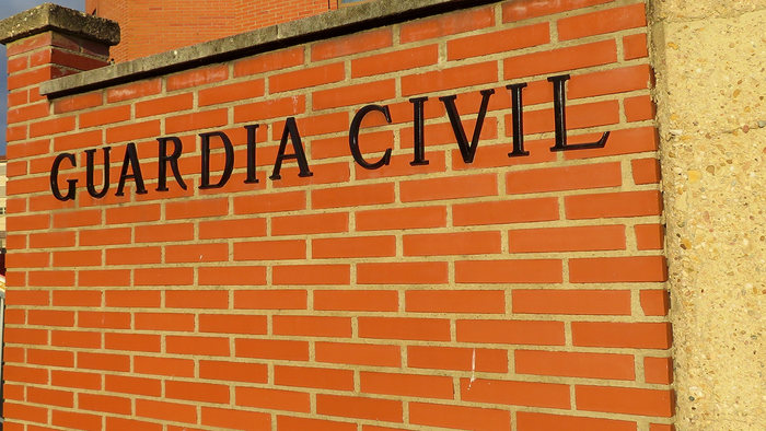 The words “Guardia Civil” in black letters on a red brick wall at the entrance to a brick building, with a raised barrier and street lamps to the left.