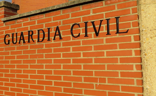 The words “Guardia Civil” in black letters on a red brick wall at the entrance to a brick building, with a raised barrier and street lamps to the left.