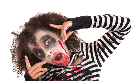 Child with curly hair in zombie makeup, with dark eye shadow and fake blood around the mouth, wearing a black and white striped top and posing against a white background.