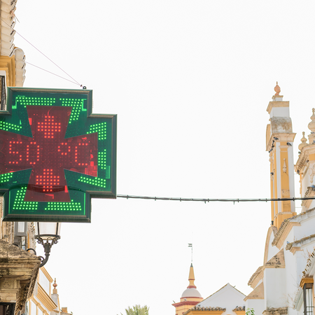 A green and red LED pharmacy cross sign on a white building displaying 50°C, above a narrow street of whitewashed buildings with yellow trim and iron balconies.
