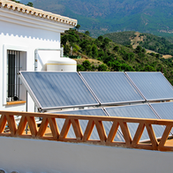 Rows of flat solar panels on a white house’s tiled terrace, behind an orange brick railing, with green forested mountains in the background.