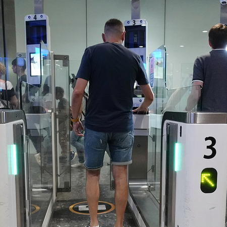 Two men seen from behind walking through automated passport gates at an airport, with green arrows lit on the gates numbered 3 and 4