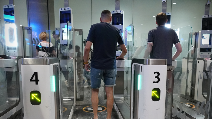 Two men seen from behind walking through automated passport gates at an airport, with green arrows lit on the gates numbered 3 and 4