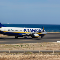 A white and blue Ryanair Boeing 737 on an airport runway beside the sea, with a fence in the foreground and a coastal town in the distance under a blue sky