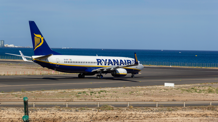 A white and blue Ryanair Boeing 737 on an airport runway beside the sea, with a fence in the foreground and a coastal town in the distance under a blue sky