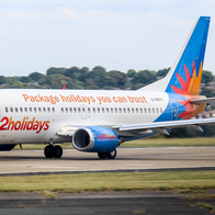 A white Jet2holidays Boeing 737 with a blue tail taxiing along a runway, with trees and houses in the background.
