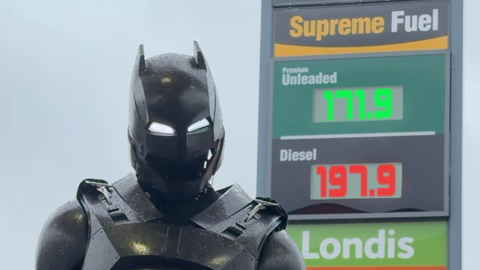 A figure in a black armoured Batman-style suit and helmet with glowing white eyes stands in the rain in front of a petrol station price sign showing unleaded at 171.9 and diesel at 197.9.