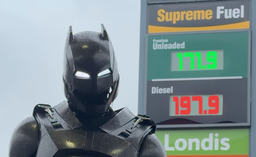 A figure in a black armoured Batman-style suit and helmet with glowing white eyes stands in the rain in front of a petrol station price sign showing unleaded at 171.9 and diesel at 197.9.