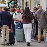 A crowd of people with suitcases and bags waiting on a pavement outside a cream-coloured apartment building on a tree-lined street.