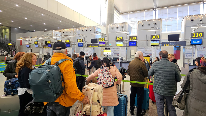 Travellers with backpacks and suitcases queue at a row of numbered check-in desks in an airport terminal.