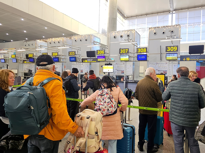 Travellers with backpacks and suitcases queue at a row of numbered check-in desks in an airport terminal.