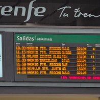 Orange LED departures board with Adif and Renfe branding, listing trains to Madrid Atocha, Sevilla Santa Justa and Málaga, next to a large wall clock.