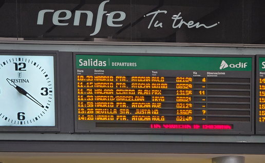 Orange LED departures board with Adif and Renfe branding, listing trains to Madrid Atocha, Sevilla Santa Justa and Málaga, next to a large wall clock.