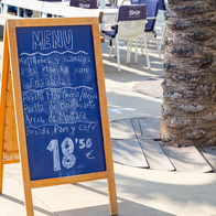 A chalkboard menu on a wooden A-frame stand outside a restaurant terrace, listing grilled mussels and prawns, paella and rice dishes, and a drink, bread and coffee for 18.50 euros.