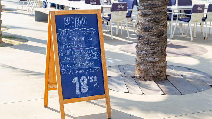 A chalkboard menu on a wooden A-frame stand outside a restaurant terrace, listing grilled mussels and prawns, paella and rice dishes, and a drink, bread and coffee for 18.50 euros.