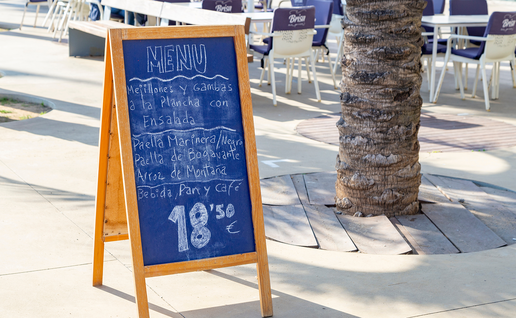 A chalkboard menu on a wooden A-frame stand outside a restaurant terrace, listing grilled mussels and prawns, paella and rice dishes, and a drink, bread and coffee for 18.50 euros.