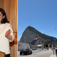 A dark-haired woman in a cream top speaks at a wooden lectern with two microphones in a gilded room, next to a view of the Rock of Gibraltar rising behind a road and border buildings, with a Spanish flag flying in the foreground.