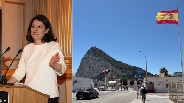A dark-haired woman in a cream top speaks at a wooden lectern with two microphones in a gilded room, next to a view of the Rock of Gibraltar rising behind a road and border buildings, with a Spanish flag flying in the foreground.