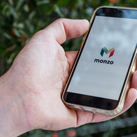 A hand holding a smartphone in a gold-coloured case, with the Monzo logo on the screen against a blurred green hedge.