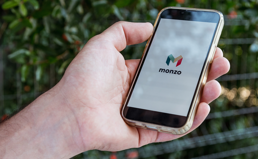 A hand holding a smartphone in a gold-coloured case, with the Monzo logo on the screen against a blurred green hedge.