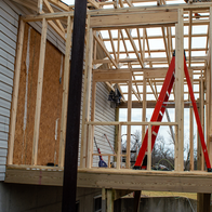 Wooden frame of a house extension with roof trusses, a red ladder and two builders working on the roof.