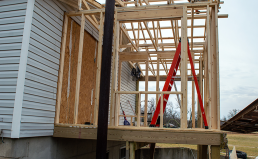 Wooden frame of a house extension with roof trusses, a red ladder and two builders working on the roof.