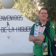 A ONCE lottery seller in a green vest holds a board of lottery tickets beside a white sign that reads ‘Bienvenidos a Fuente de la Higuera’.